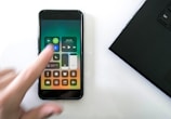 A hand blurred slightly in motion is interacting with a smartphone displaying the iPhone control center. The control center shows various icons including Wi-Fi, Bluetooth, and music controls. A laptop partially visible on the right side forms part of the scene, all placed on a white surface.