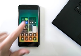 A hand blurred slightly in motion is interacting with a smartphone displaying the iPhone control center. The control center shows various icons including Wi-Fi, Bluetooth, and music controls. A laptop partially visible on the right side forms part of the scene, all placed on a white surface.