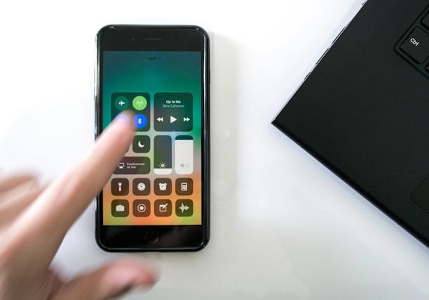 A hand blurred slightly in motion is interacting with a smartphone displaying the iPhone control center. The control center shows various icons including Wi-Fi, Bluetooth, and music controls. A laptop partially visible on the right side forms part of the scene, all placed on a white surface.