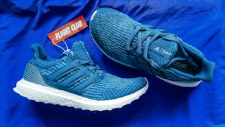 A pair of stylish blue athletic sneakers with white soles is placed on a matching blue background. One shoe is positioned upright while the other is lying on its side. A red tag with the words 'FLIGHT CLUB' is attached to the upright shoe. The sneakers exhibit a textured pattern with prominent branding elements including the Adidas logo and the label 'Parley'.