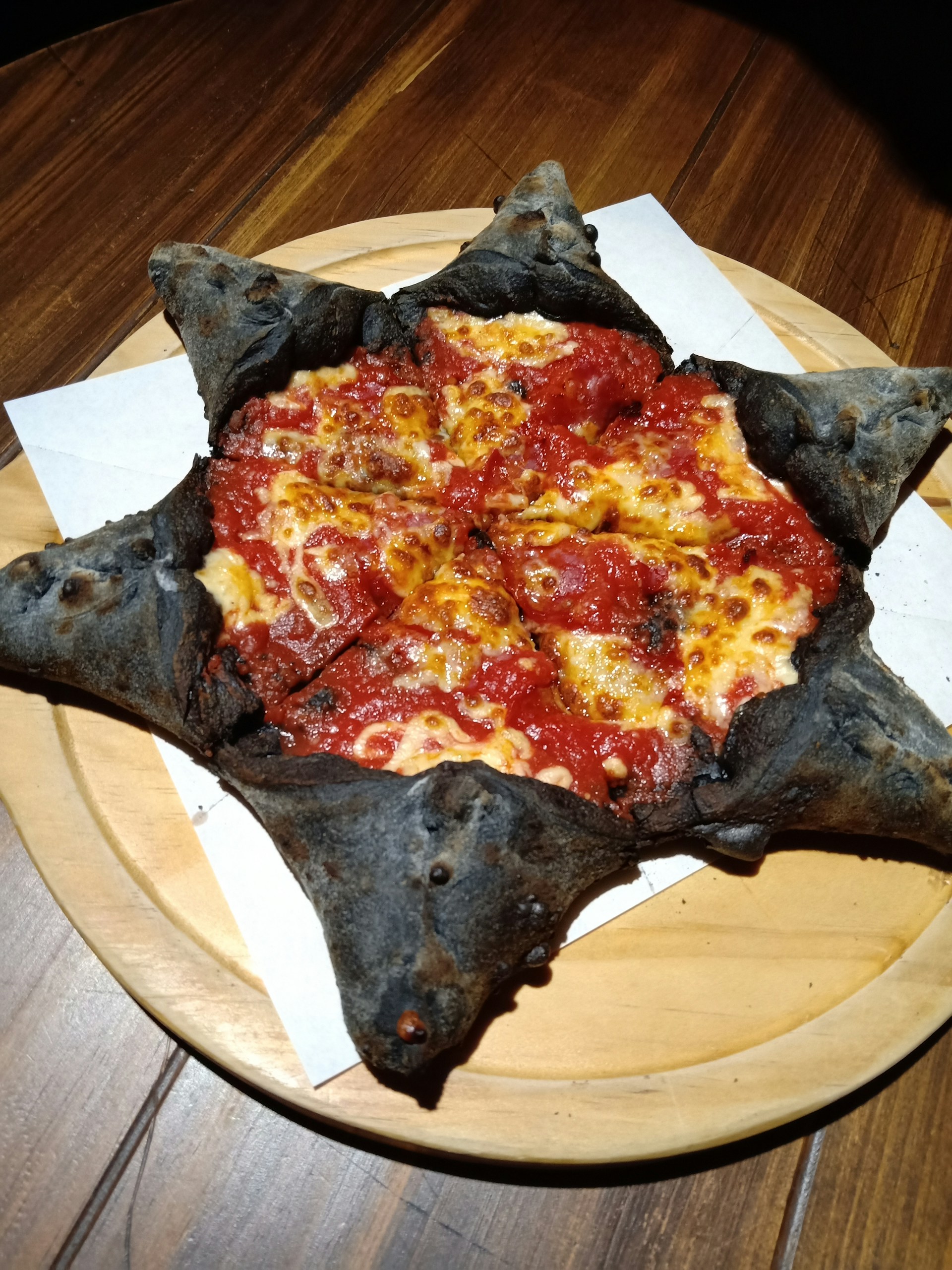 A pizza with a unique charcoal black crust shaped into pointed star-like formations. The toppings include bubbling cheese and a generous amount of tomato sauce, contributing to a rich red and yellow color contrast. It is placed on a wooden serving board against a wooden table.