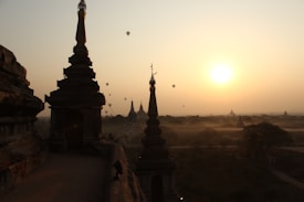 A scenic view during sunrise featuring ancient pagodas silhouetted against the bright morning sky. Several hot air balloons float in the distance, adding a sense of calm and exploration. The landscape extends into the misty horizon, creating a tranquil atmosphere.