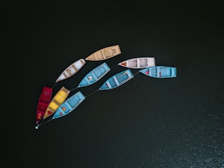 Nine small rowboats are floating on a dark green, rippled water surface. The boats are connected by ropes and vary in color, including red, blue, yellow, beige, and white. Each boat is slightly worn, suggesting regular use, and they are arranged in a rough circular formation.