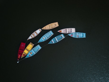 Nine small rowboats are floating on a dark green, rippled water surface. The boats are connected by ropes and vary in color, including red, blue, yellow, beige, and white. Each boat is slightly worn, suggesting regular use, and they are arranged in a rough circular formation.