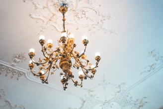 An ornate, antique chandelier with multiple light bulbs hangs from a ceiling with intricate, decorative plasterwork. The chandelier features detailed metalwork and floral motifs, casting a warm glow on the elegant surroundings.