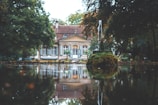 A tranquil scene featuring a house with a red and tan roof, surrounded by lush greenery. In the foreground, there is a small moss-covered fountain with a stream of water shooting upwards, creating a serene reflection on a calm body of water.