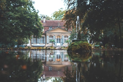 A tranquil scene featuring a house with a red and tan roof, surrounded by lush greenery. In the foreground, there is a small moss-covered fountain with a stream of water shooting upwards, creating a serene reflection on a calm body of water.