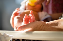 A close-up of a baby's feet resting on an adult's hand, with soft lighting creating a warm and gentle atmosphere. The scene suggests tenderness and care, capturing a moment of bonding.