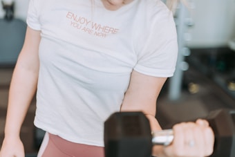 Person wearing a white t-shirt with the text 'ENJOY WHERE YOU ARE NOW' is holding a dumbbell, engaging in a bicep curl exercise. Background is blurred, suggesting an indoor setting, possibly a gym.