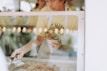 A person is seen holding a stack of cookies, wearing a light-colored apron inside a bakery setting. The focus is on the cookies, while the background is softly blurred with warm lighting. The scene is viewed through a glass window.