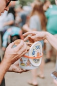 Two people engaged in sharing or passing cookies from a colorful cookie jar with cartoon illustrations. The background is blurred, showing an outdoor gathering with other people present.