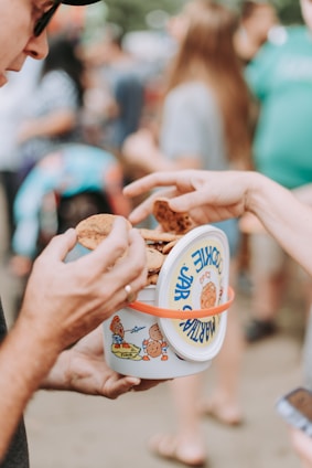 Two people engaged in sharing or passing cookies from a colorful cookie jar with cartoon illustrations. The background is blurred, showing an outdoor gathering with other people present.