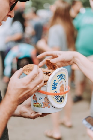 Two people engaged in sharing or passing cookies from a colorful cookie jar with cartoon illustrations. The background is blurred, showing an outdoor gathering with other people present.