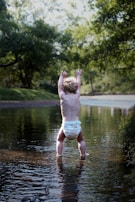 A toddler wearing a diaper stands in a shallow stream surrounded by lush greenery. The child faces away and raises their arms, possibly playing or reaching out. Sunlight filters through the trees, casting dappled shadows on the water.