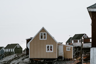 A series of small, wooden houses with steep roofs are situated on a rocky landscape. The main house in the foreground is beige with white window frames and has a triangular roof. Other houses in the background are darker in color and have similar architectural styles. The scene is set against a clear sky.