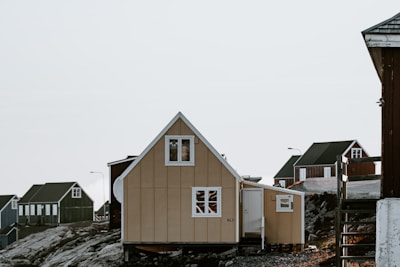 A series of small, wooden houses with steep roofs are situated on a rocky landscape. The main house in the foreground is beige with white window frames and has a triangular roof. Other houses in the background are darker in color and have similar architectural styles. The scene is set against a clear sky.