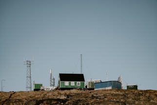 A remote station with several green and blue buildings sits atop a rocky hill. Various antennas and communication towers are positioned around the structures, indicating a focus on telecommunications or research. The sky is clear, and there is no vegetation around the buildings.