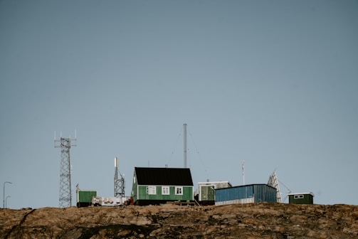 A remote station with several green and blue buildings sits atop a rocky hill. Various antennas and communication towers are positioned around the structures, indicating a focus on telecommunications or research. The sky is clear, and there is no vegetation around the buildings.