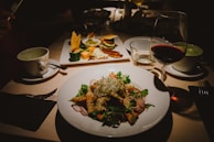 A dimly lit restaurant table set with a variety of food and drink items. The focus is on a white plate filled with a fresh salad, including greens, radishes, and croutons. A glass of red wine is visible to the right, alongside a small white coffee cup. In the background, there is a platter with cheese, crackers, and small dishes containing dips or condiments.