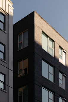 A modern urban building with a dark facade stands adjacent to a lighter, classic building. The architecture displays a mixture of contemporary and traditional styles, with sunlight casting sharp shadows across the structures. Windows on both buildings reflect a clear sky, creating a sharp contrast between light and shadow.