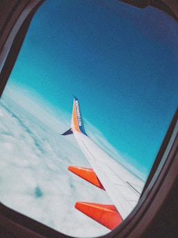 An airplane wing with orange accents is seen from a passenger window, soaring high above a blanket of clouds, under a clear blue sky. The tail fin displays branding.