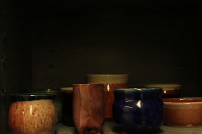 The image depicts a collection of pottery pieces placed on a shelf. The pottery includes several ceramic cups and bowls of varying shapes and colors, with a dark background creating a moody ambiance. The ceramics feature glazes in tones of white, pink, blue, and brown, showcasing different textures and finishes.