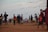 A lively beach scene with people of various ages walking and flying kites. The sky is filled with colorful kites, and the ground is sandy. Some individuals are holding kites, while others are engaged in conversations or strolling.