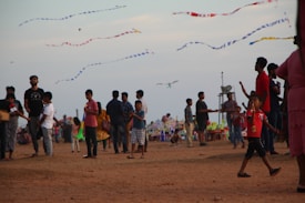 A lively beach scene with people of various ages walking and flying kites. The sky is filled with colorful kites, and the ground is sandy. Some individuals are holding kites, while others are engaged in conversations or strolling.