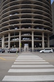 A multi-story circular parking garage with vehicles parked on various levels is shown. The architecture features open spaces on each level revealing the parked cars from the side view. A street with a zebra crossing and several vehicles, including cars and vans, is visible in front of the parking structure.