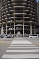 A multi-story circular parking garage with vehicles parked on various levels is shown. The architecture features open spaces on each level revealing the parked cars from the side view. A street with a zebra crossing and several vehicles, including cars and vans, is visible in front of the parking structure.
