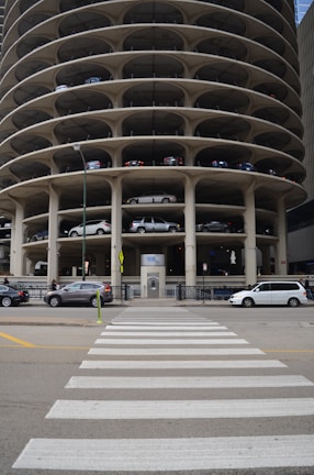 A multi-story circular parking garage with vehicles parked on various levels is shown. The architecture features open spaces on each level revealing the parked cars from the side view. A street with a zebra crossing and several vehicles, including cars and vans, is visible in front of the parking structure.