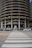 A multi-story circular parking garage with vehicles parked on various levels is shown. The architecture features open spaces on each level revealing the parked cars from the side view. A street with a zebra crossing and several vehicles, including cars and vans, is visible in front of the parking structure.