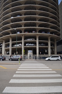 A multi-story circular parking garage with vehicles parked on various levels is shown. The architecture features open spaces on each level revealing the parked cars from the side view. A street with a zebra crossing and several vehicles, including cars and vans, is visible in front of the parking structure.