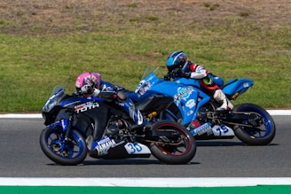 Two motorcyclists are racing side by side on a track. One rider is on a black and blue bike with the number 30 and is wearing a pink helmet. The other rider is on a blue motorcycle with the number 79 and has a blue helmet. The track is bordered by areas of grass.
