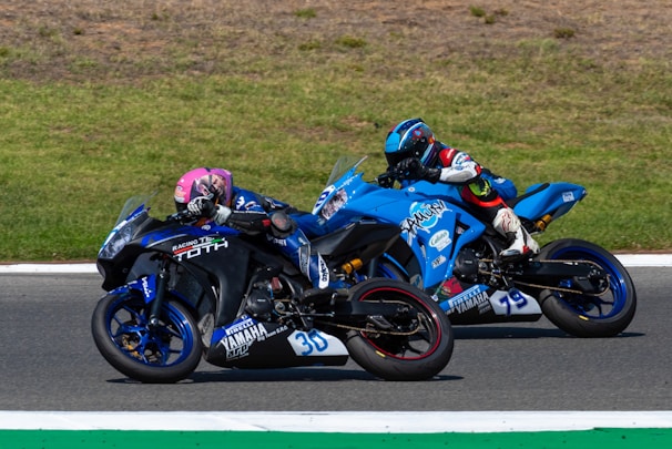 Two motorcyclists are racing side by side on a track. One rider is on a black and blue bike with the number 30 and is wearing a pink helmet. The other rider is on a blue motorcycle with the number 79 and has a blue helmet. The track is bordered by areas of grass.