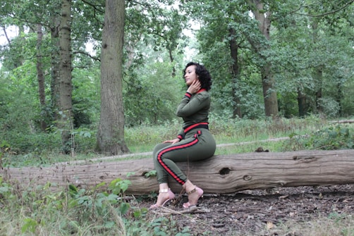 A woman in a green tracksuit with red accents sits on a large fallen tree trunk in a forest setting. She is surrounded by lush greenery, including tall trees and abundant undergrowth. Her gaze is thoughtful, and she appears to be enjoying the tranquil outdoor environment.