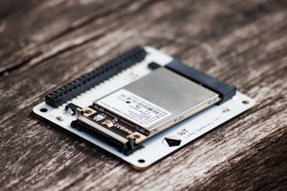 A close-up of an electronic circuit board featuring a metallic component mounted on a white base, with visible connectors and pins. The board is placed on a textured wooden surface.