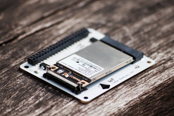 A close-up of an electronic circuit board featuring a metallic component mounted on a white base, with visible connectors and pins. The board is placed on a textured wooden surface.