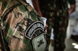 A detailed view of a camouflage military uniform with a patch that reads 'Operações com Cães'. The background shows another individual wearing a similar uniform, holding a white cup.