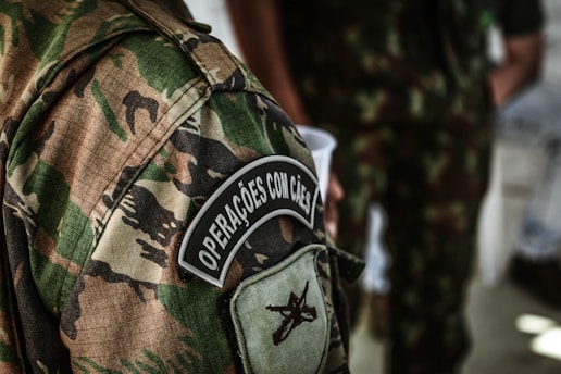A detailed view of a camouflage military uniform with a patch that reads 'Operações com Cães'. The background shows another individual wearing a similar uniform, holding a white cup.