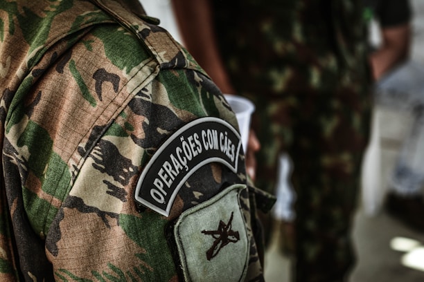 A detailed view of a camouflage military uniform with a patch that reads 'Operações com Cães'. The background shows another individual wearing a similar uniform, holding a white cup.