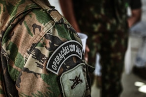 A detailed view of a camouflage military uniform with a patch that reads 'Operações com Cães'. The background shows another individual wearing a similar uniform, holding a white cup.