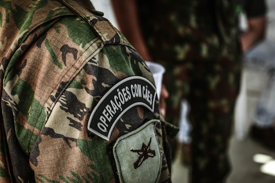 A detailed view of a camouflage military uniform with a patch that reads 'Operações com Cães'. The background shows another individual wearing a similar uniform, holding a white cup.