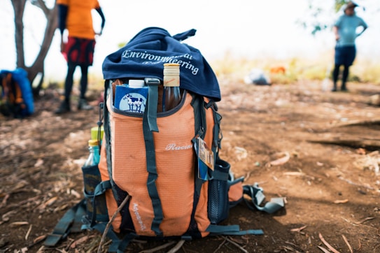 A large orange and black backpack rests on a dirt trail with various items protruding from its pockets, including a bottle and a piece of clothing with the word 'Engineering' visible. In the background, a couple of people are standing on the trail, with trees and foliage scattered around.