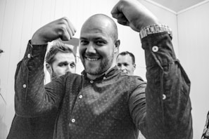 A group of three men are gathered in a room with a man in the foreground smiling and flexing his muscles. Another man stands closely behind him, partially obscured, and a third man is in the background, looking towards the foreground. The room has light-colored walls and a high ceiling. The overall setting appears casual.