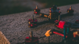 A close-up of a drone frame rests on a textured concrete surface. The drone has prominent red and black motor components with metallic bolts visible on the top. The focus is on the mechanical elements, providing a detailed view of the drone's construction.