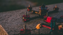 A close-up of a drone frame rests on a textured concrete surface. The drone has prominent red and black motor components with metallic bolts visible on the top. The focus is on the mechanical elements, providing a detailed view of the drone's construction.