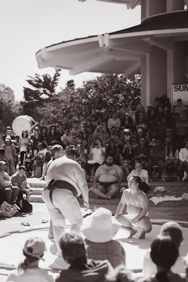Two sumo wrestlers are engaged in a match on a raised platform surrounded by a large crowd of spectators. The outdoor setting includes a backdrop of trees and a structure with a canopy roof. The audience appears engaged, with some people taking photos.