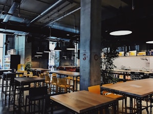 A modern industrial-style cafe with exposed ceilings and concrete pillars. There are several wooden tables and yellow chairs neatly arranged throughout the space. Hanging lights provide illumination, and plants are positioned strategically around the room. In the background, there is a tiled counter area with menus written on blackboards.