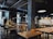 A modern industrial-style cafe with exposed ceilings and concrete pillars. There are several wooden tables and yellow chairs neatly arranged throughout the space. Hanging lights provide illumination, and plants are positioned strategically around the room. In the background, there is a tiled counter area with menus written on blackboards.